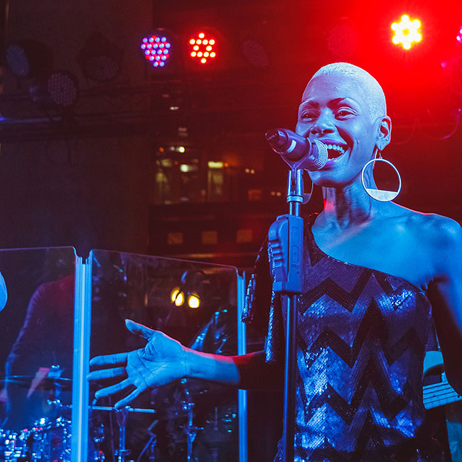 Female vocalist behind microphone singing with live band in background.